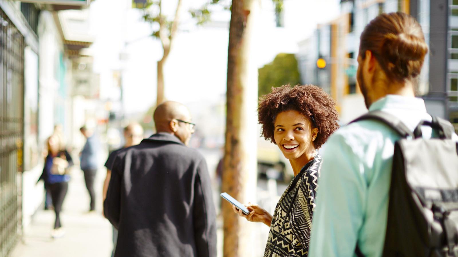 Woman holding a mobile phone, smiling at a person behind her.